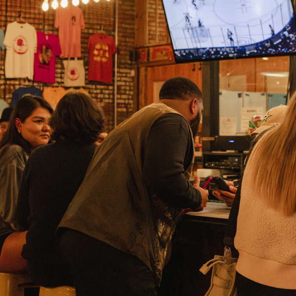 Guests ordering at the bar inside Four Corners Brewing Co. taproom with merch wall and hockey game on TV
