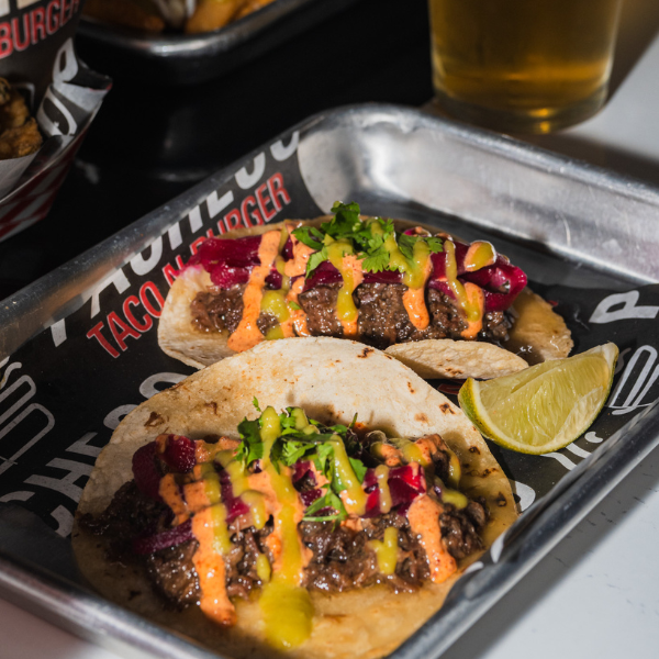 Two street tacos topped with spicy aioli, pickled beets, and cilantro on a Pacheco Taco N Burger tray with a beer in the background