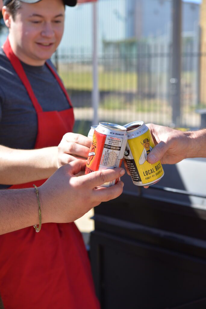 A smiling person in a red apron clinks open cans of Four Corners Brewing Co. Local Buzz Honey-Rye Golden Ale and El Chingón with two other people outdoors.