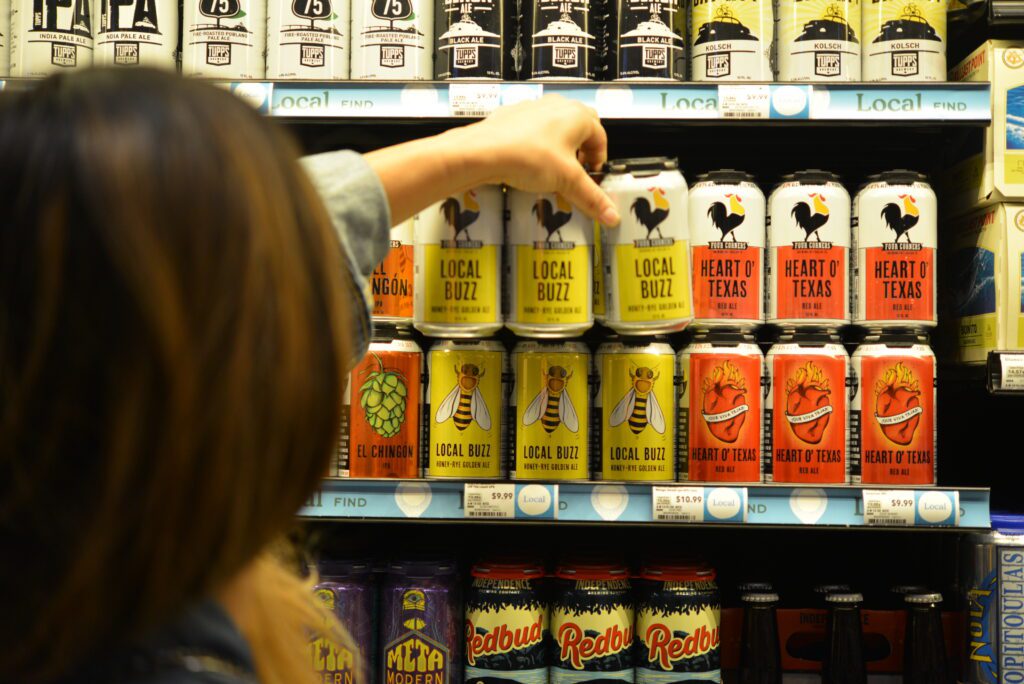 A shopper reaches for a can of Four Corners Brewing Co. Local Buzz from a well-stocked retail beer shelf featuring Local Buzz, Heart O' Texas, and El Chingón, tagged under a "Local Find" shelf label.