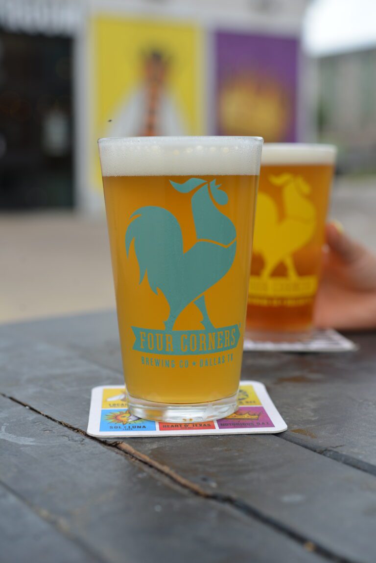 A full Four Corners Brewing Co. branded pint glass of golden beer on a lotería-style coaster on a weathered wood table, with a second pint glass being held in the background.