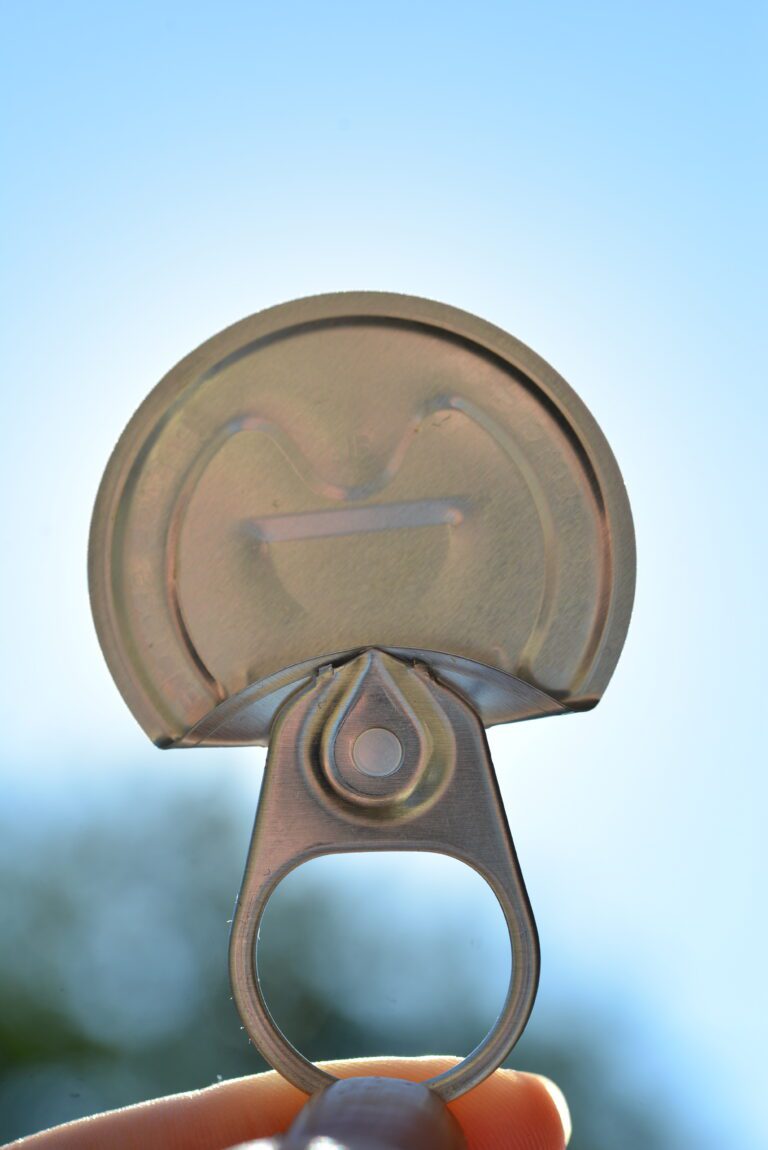 Close-up of a fully removed 360° pop top beer can lid held up against a clear blue sky, with the pull tab and ring visible beneath it.