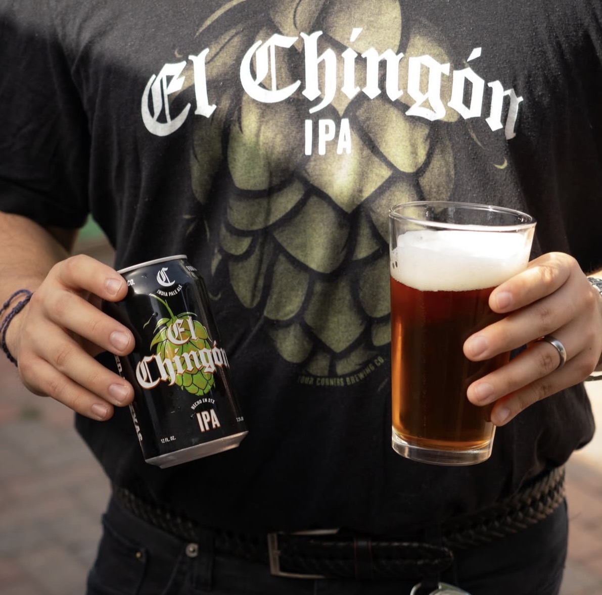 A person wearing a Four Corners Brewing Co. El Chingón IPA t-shirt holds a can of El Chingón in one hand and a full pint glass of the amber IPA in the other.