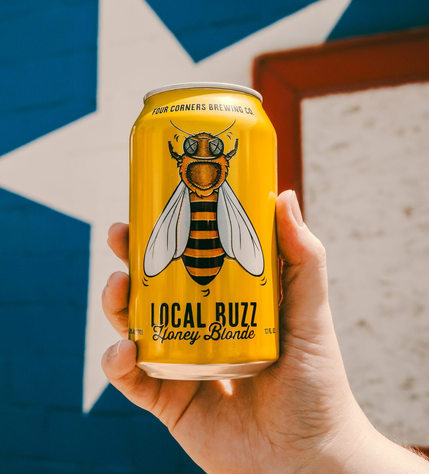 A hand holds up a can of Four Corners Brewing Co. Local Buzz Honey Blonde against a blue wall painted with a large white Lone Star.