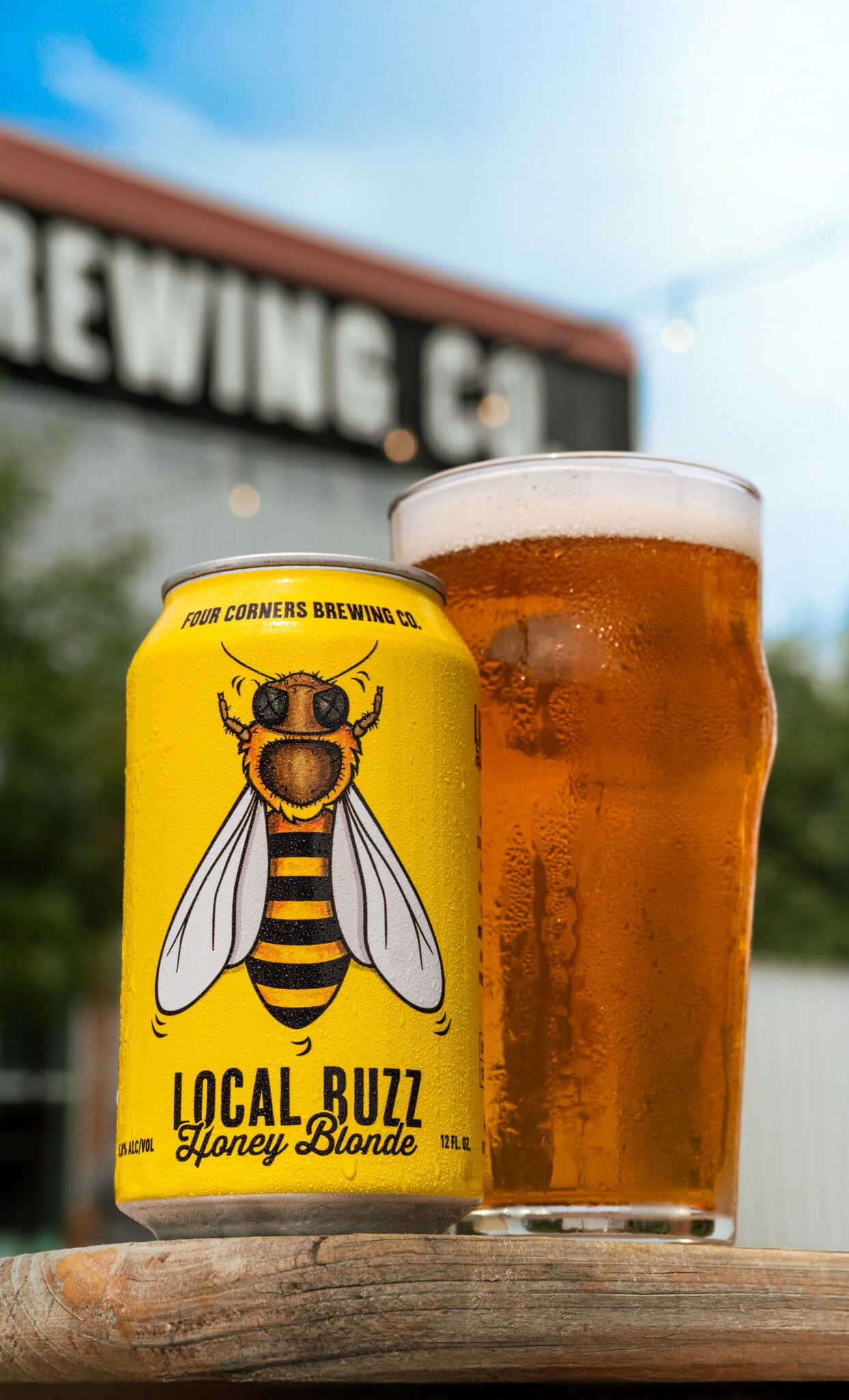 A can of Four Corners Brewing Co. Local Buzz Honey Blonde beside a cold, condensation-covered pint glass of golden ale on a weathered wood surface, with the brewery building visible in the background.
