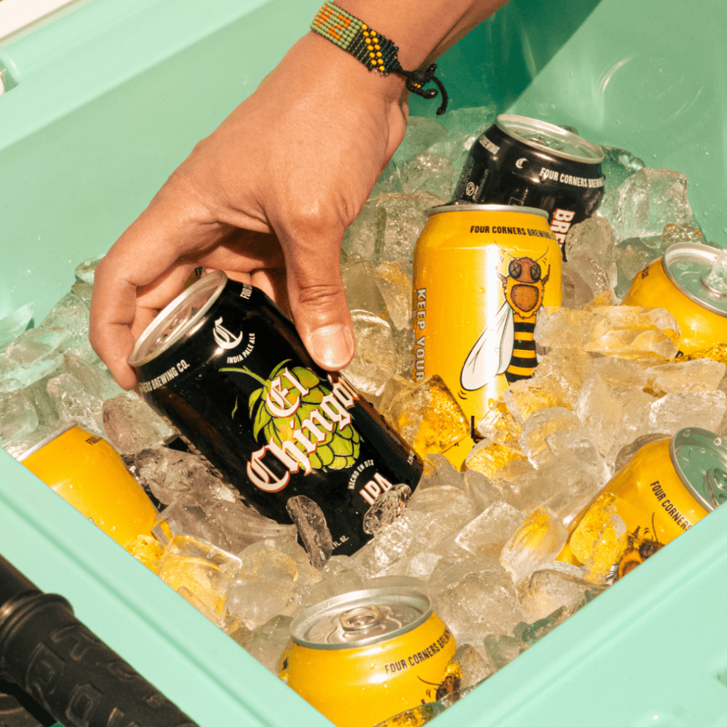 Hand with beaded bracelet reaching into a mint green cooler packed with El Chingón IPA and Local Buzz Honey Blonde cans on ice