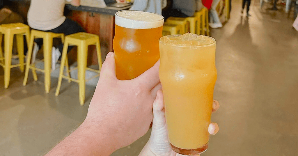 Two hands toasting a hazy IPA and a citrus beer inside Four Corners Brewing Co. taproom with yellow bar stools in background