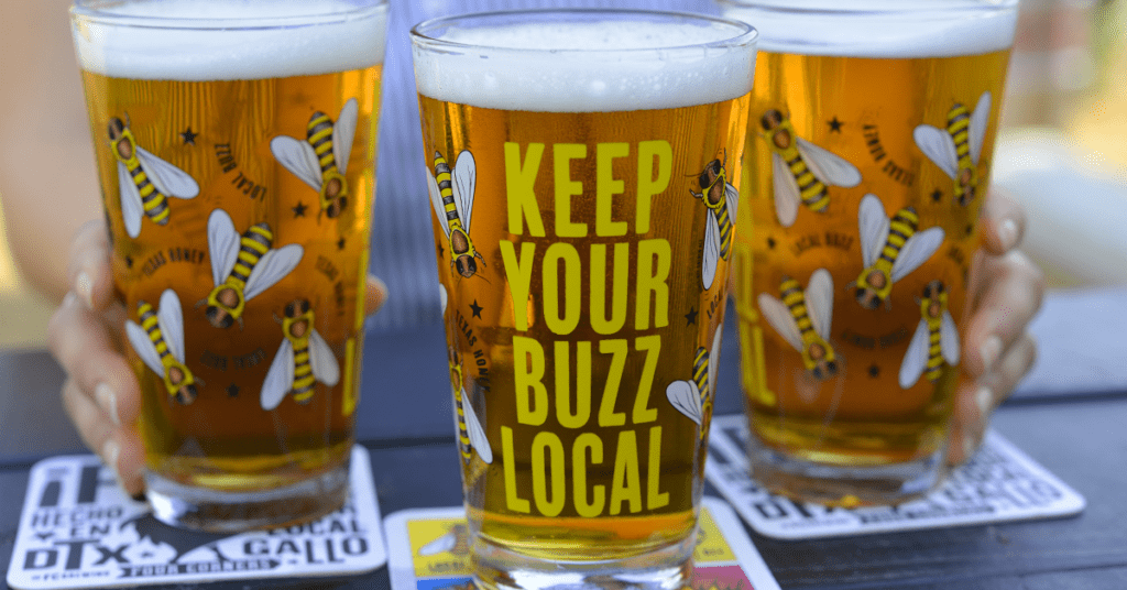 Three Four Corners Brewing Co. pint glasses printed with Keep Your Buzz Local and bee illustrations, filled with golden Local Buzz Honey Blonde
