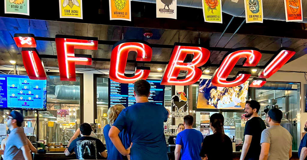 Guests ordering at the bar inside Four Corners Brewing Co. taproom beneath a large red neon FCBC sign and beer banner decorations