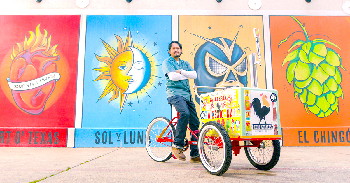 Man standing with a Paletería a la Mexicana cart in front of Four Corners Brewing Co.'s exterior mural wall featuring Que Viva Tejas, Sol y Luna, and El Chingón panels