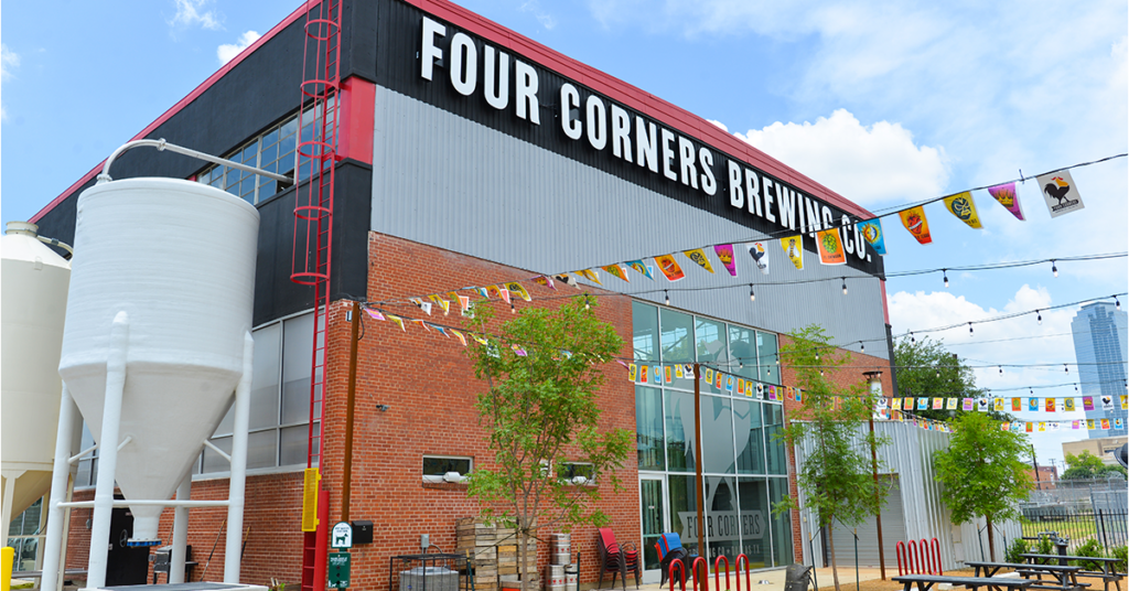 Exterior of Four Corners Brewing Co. taproom in Dallas with brick facade, grain silo, colorful papel picado banners, and Dallas skyline in background