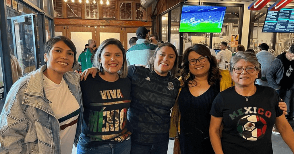 Group of five friends in Mexico soccer gear smiling at a Four Corners Brewing Co. taproom watch party with a soccer match on TV in the background