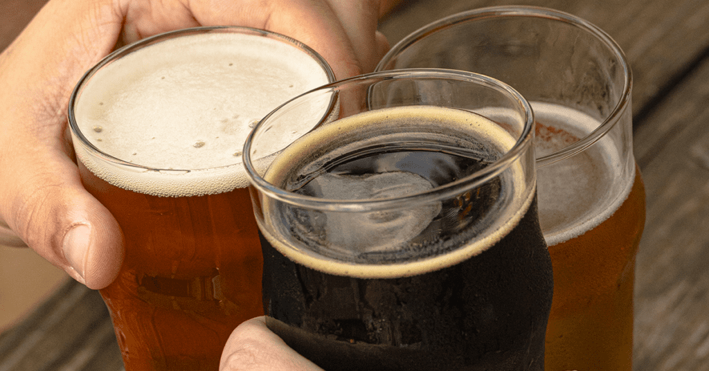 Three hands clinking pints of pale ale, stout, and amber beer outdoors