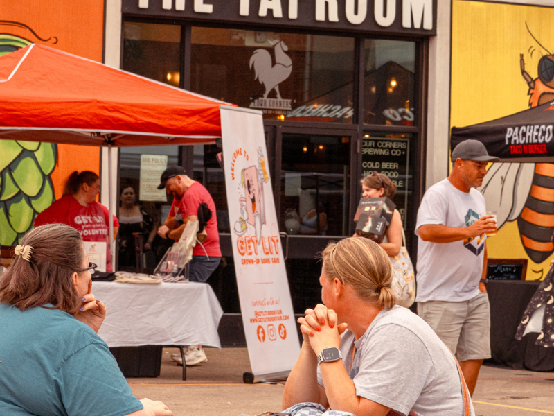 Two women sit in the foreground at an outdoor event in front of Four Corners Brewing Co.'s taproom, with a "Get Lit Grown-Up Book Fair" banner, vendor tents, and a colorful hop mural visible behind them.