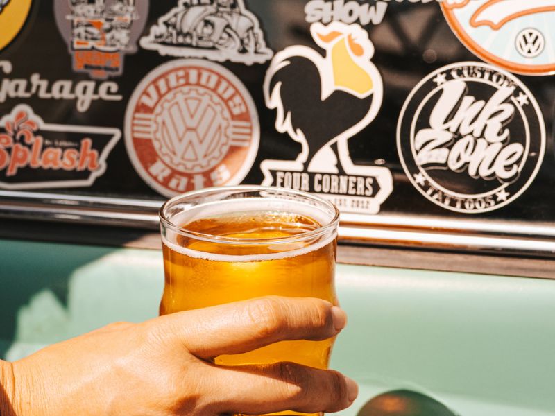 A hand holds a short glass of golden craft beer in front of a vintage car door plastered with stickers, including a Four Corners Brewing Co. rooster logo sticker alongside Volkswagen, Vicious Rods, Custom Ink Zone, and other car culture decals.
