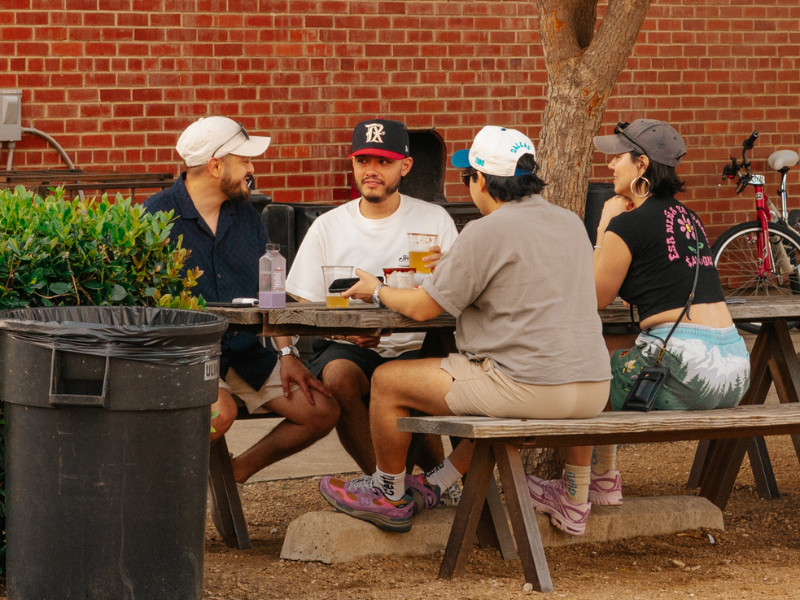 Four friends wearing baseball caps sit around a wooden picnic table against a brick wall on the Four Corners Brewing Co. outdoor patio, with pints of beer on the table and a bicycle leaning against the wall behind them.