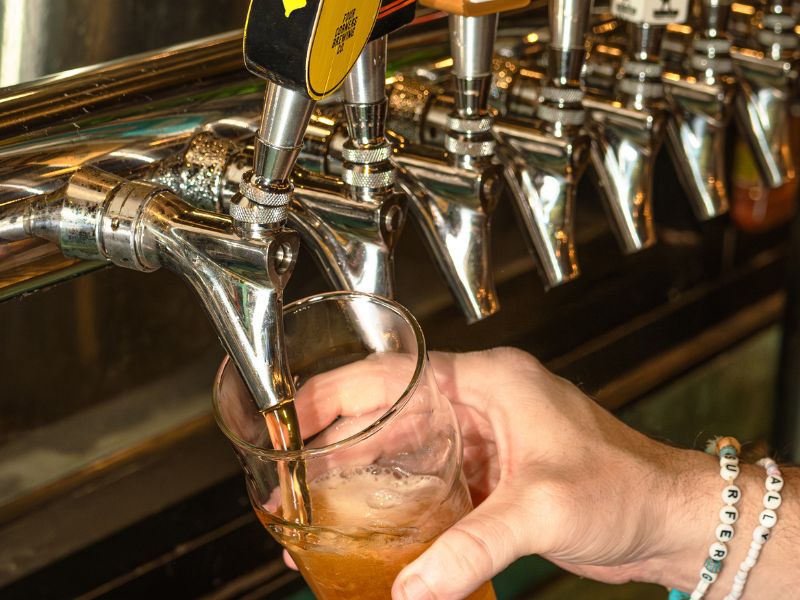A bartender's hand tilts a pint glass under a chrome draft tap handle branded with the Four Corners Brewing Co. logo, filling it with a hazy golden beer, with a row of additional taps visible in the background.