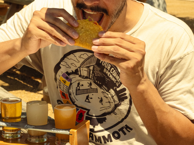 A man wearing a Four Corners Brewing Co. "Ghost Mammoth" graphic tee takes a big bite of a taco while a wooden beer flight paddle with labeled sample glasses sits on the table in front of him.