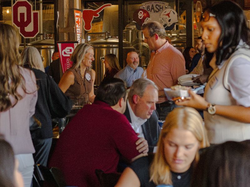 A lively crowd of adults mingles inside Four Corners Brewing Co. during a college football pre-game bash, with OU and Texas Longhorns pennants and signage decorating the taproom and brewery tanks visible in the background.